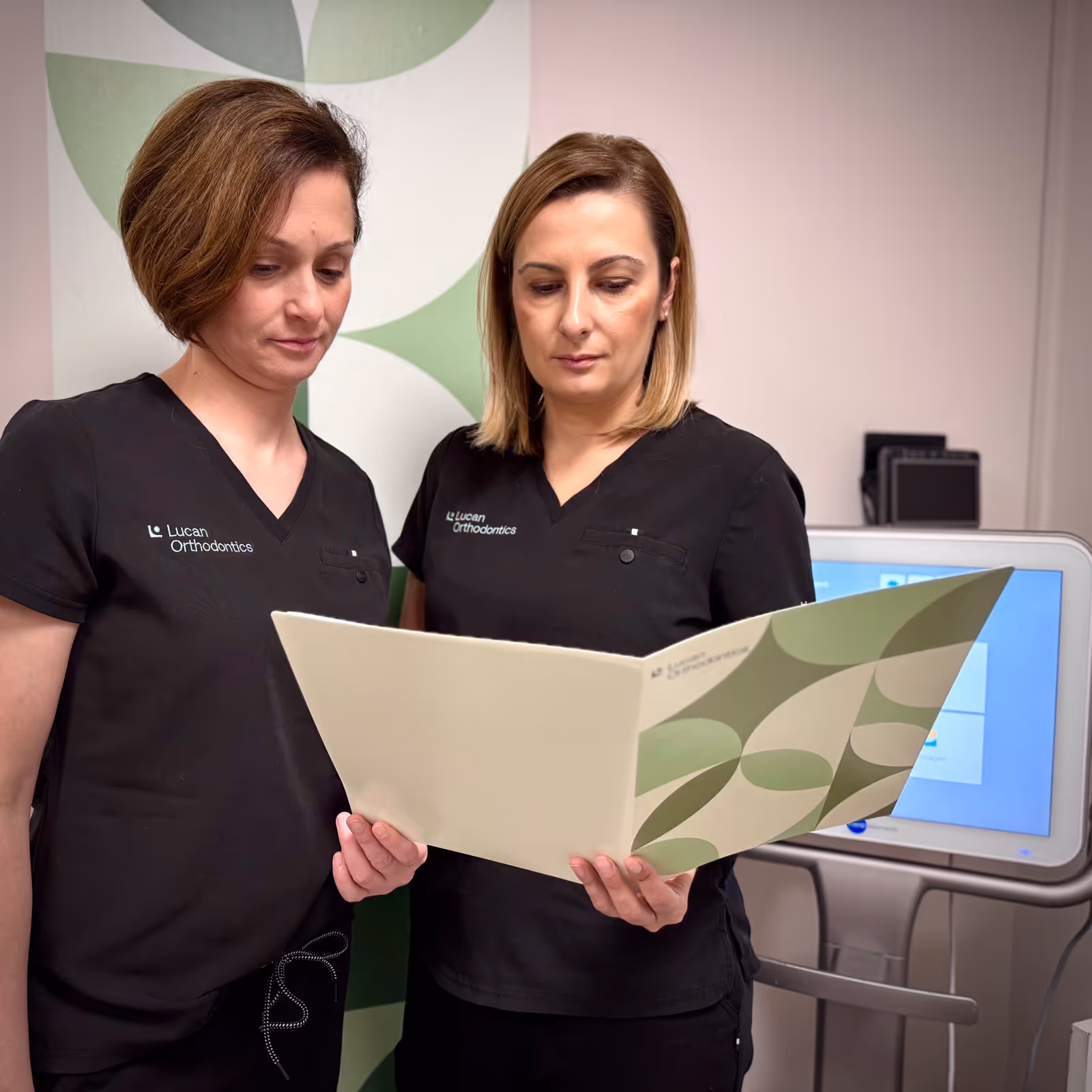 Two female orthodontic professionals in black Lucan Orthodontics scrubs reviewing a folder in a clinical setting.