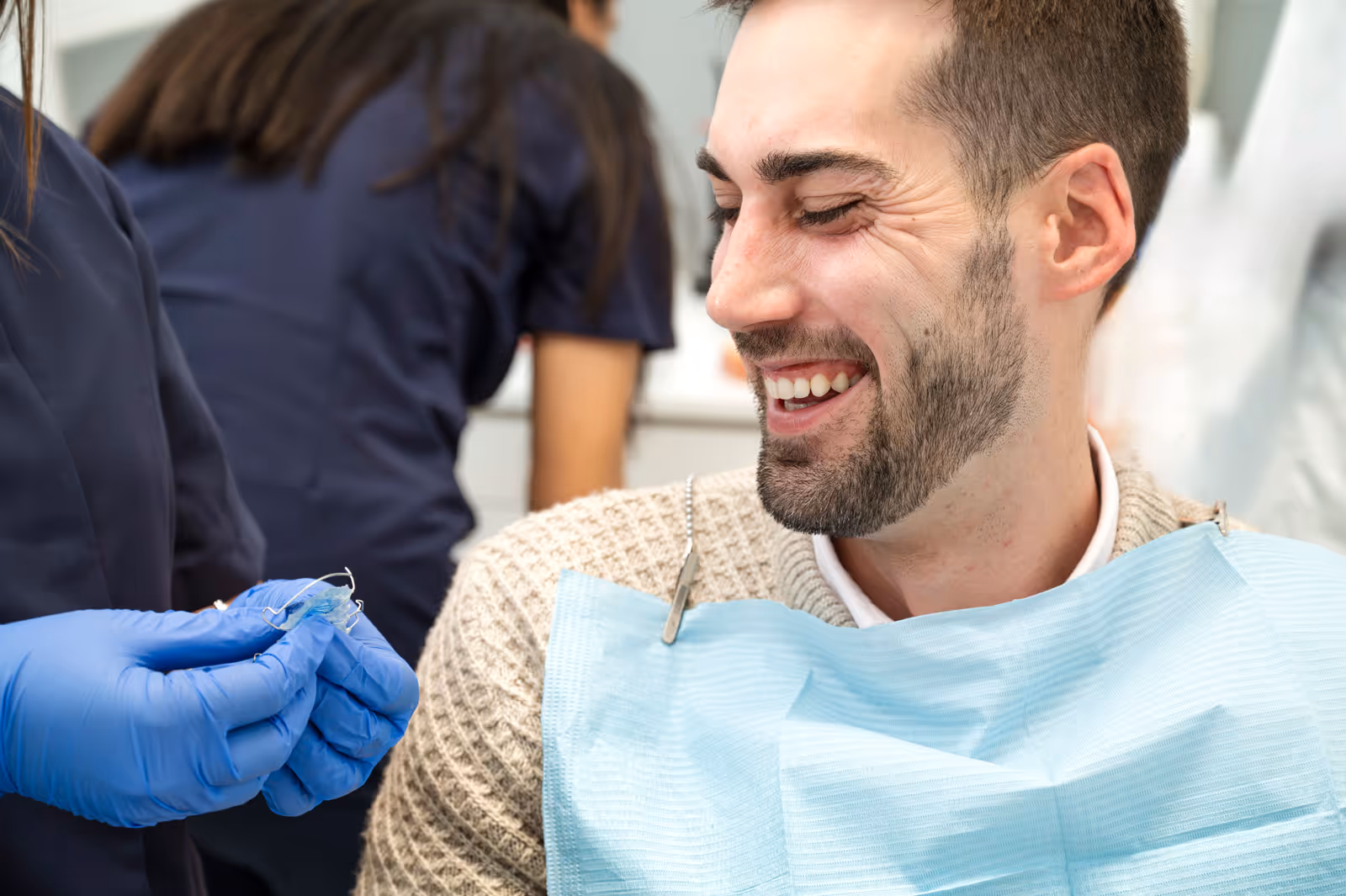 Smiling man wearing a beige sweater and blue dental bib looking at dental tools held by a gloved hand.