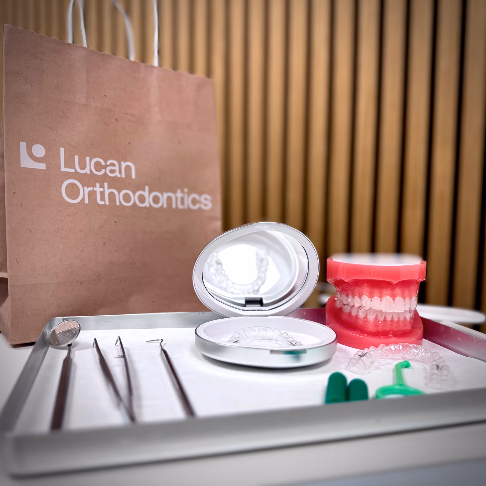 Orthodontic tools on a tray with clear aligners in a silver case and a dental model of teeth wearing braces, next to a brown paper bag labeled Lucan Orthodontics.