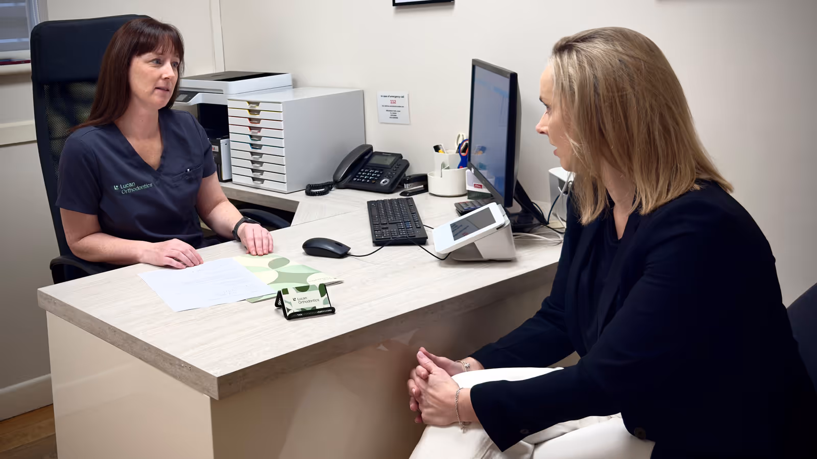 Dentist in purple scrubs showing a patient an X-ray of teeth on a monitor in a dental office.