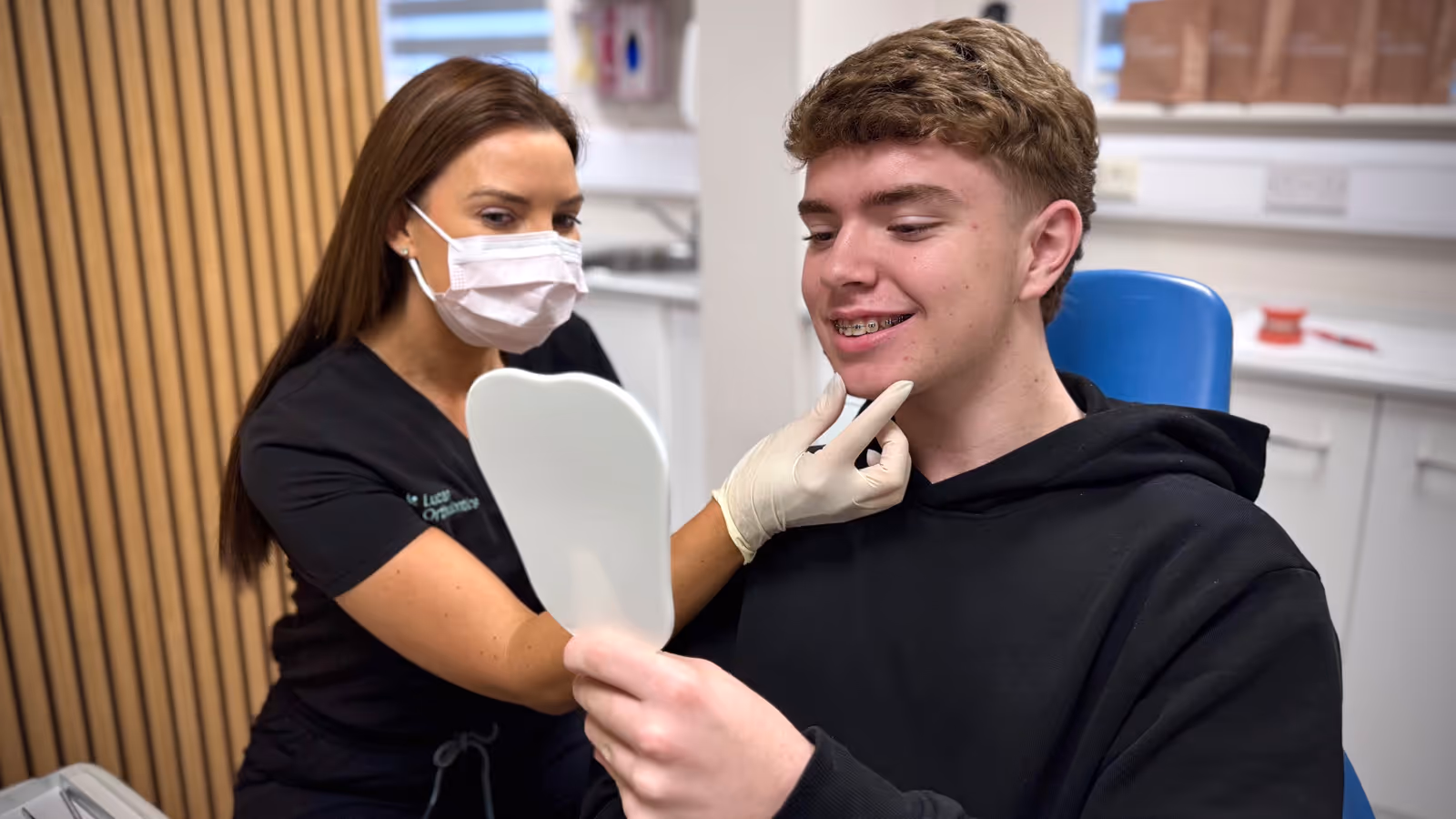 Orthodontist wearing a mask and gloves examining a male patient with braces who is looking at his teeth in a handheld mirror.