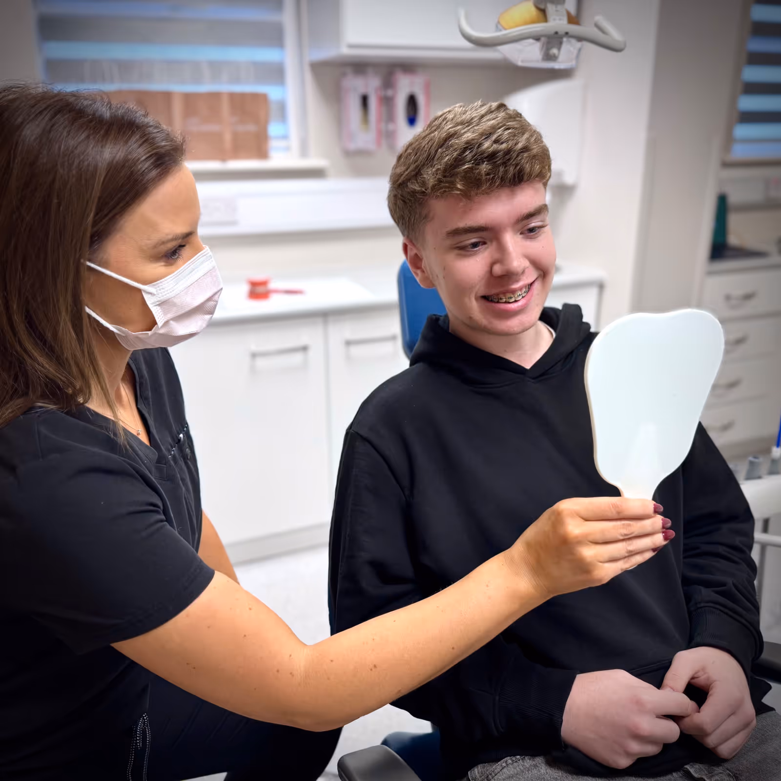 Female dentist wearing a mask shows a young male patient with braces his teeth using a hand mirror in a dental clinic.