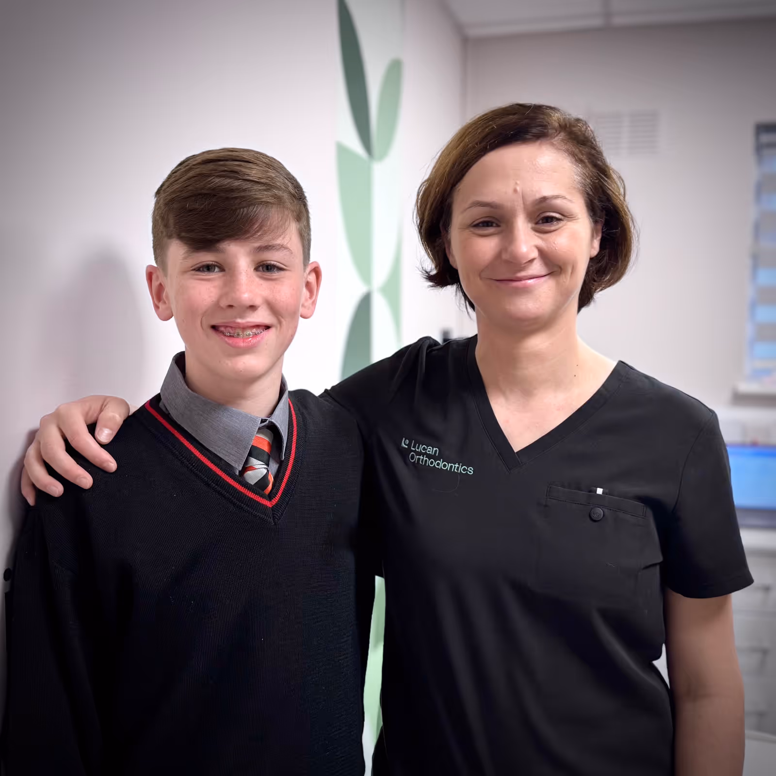 Smiling boy with braces standing next to a woman in a black Lucan Orthodontics uniform, indoors.