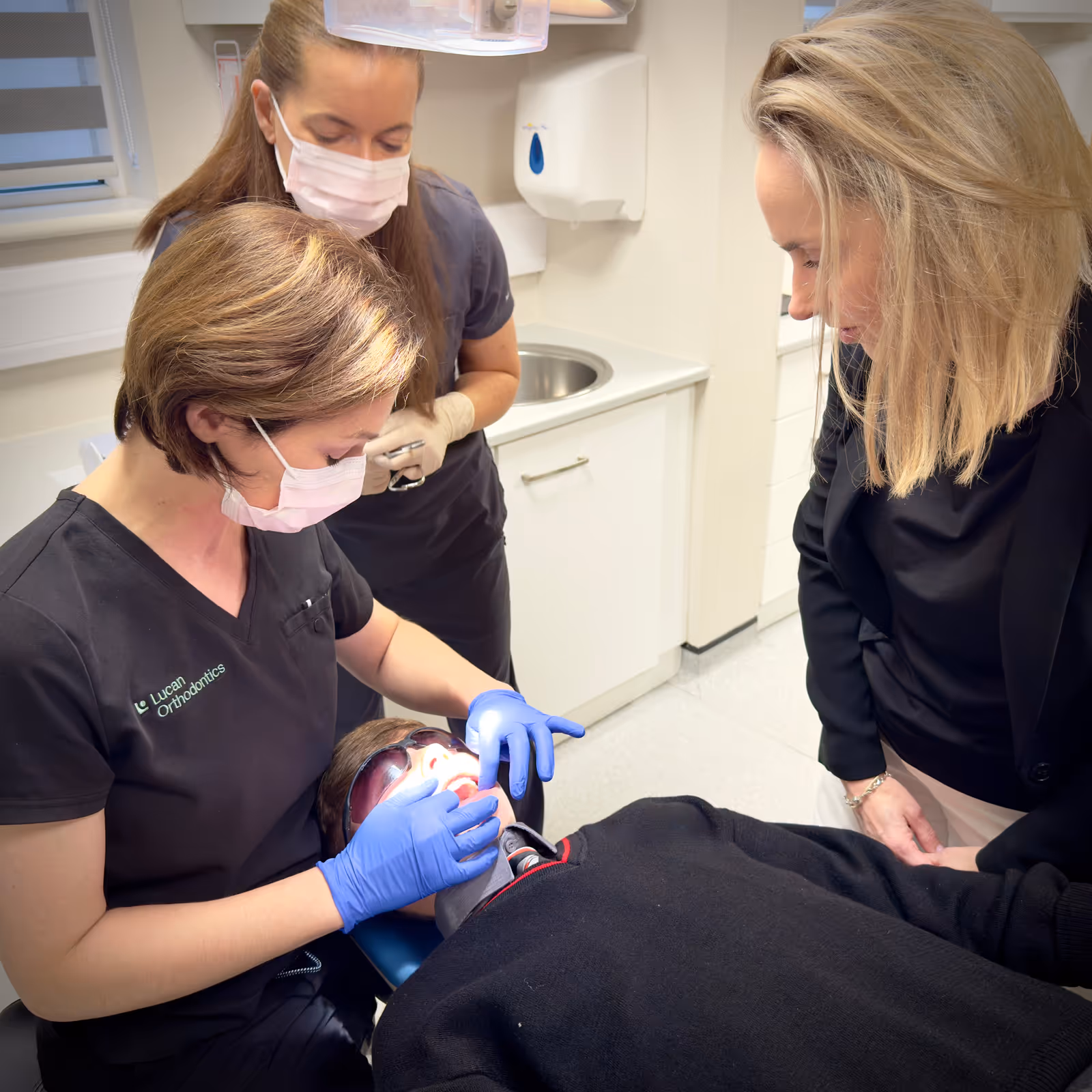 Orthodontist wearing gloves and mask examines a patient's teeth while another masked assistant and a woman observe in a dental clinic.