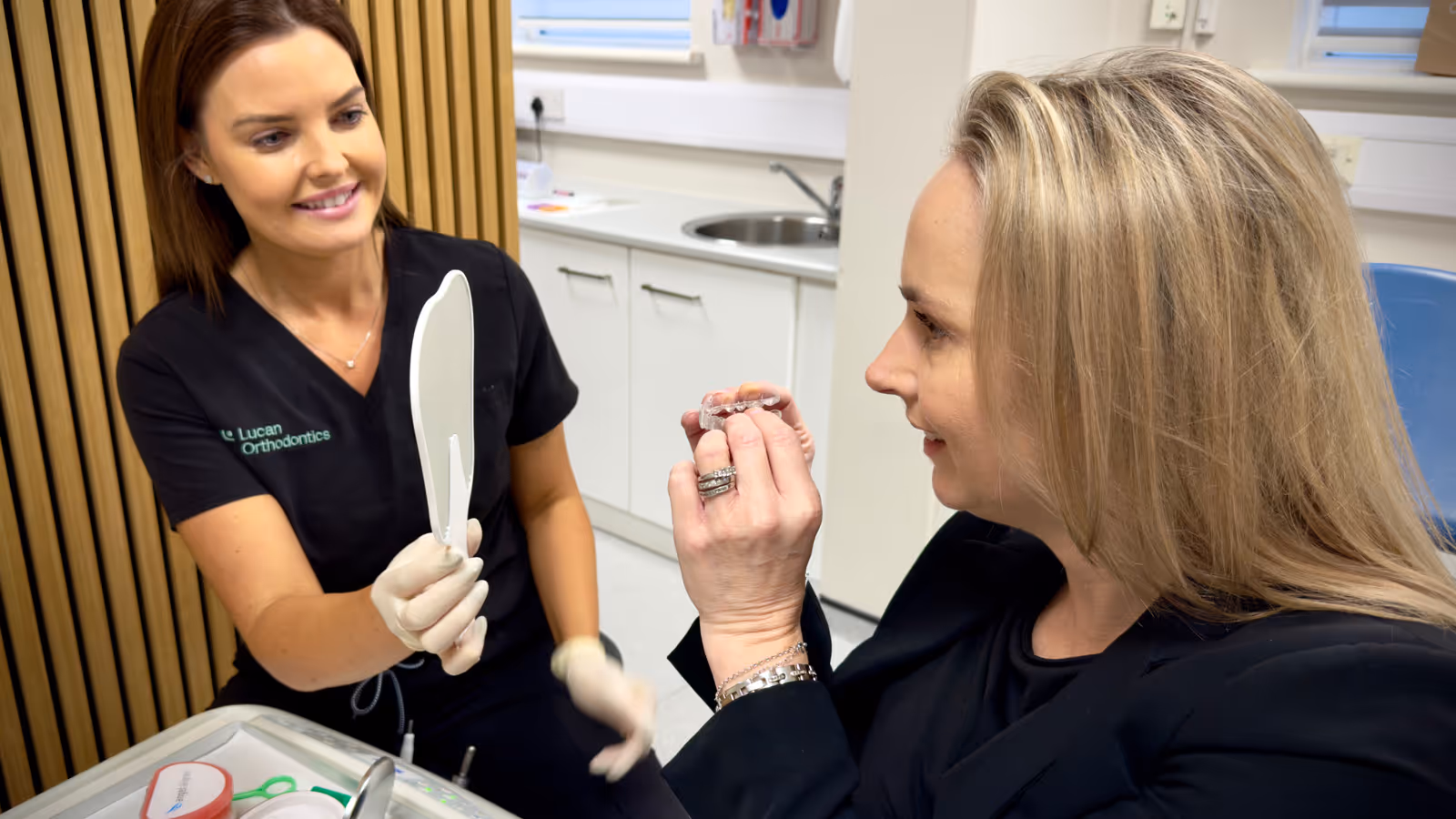 Orthodontist nurse in black scrubs holding a mirror while a patient holds a clear dental aligner in a clinic.