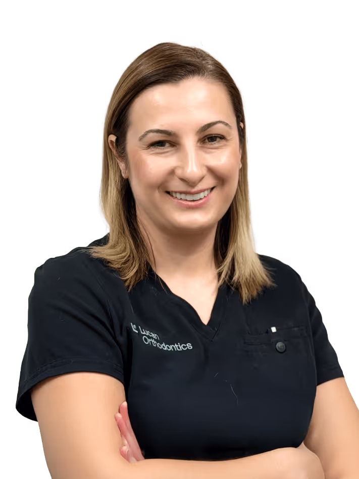 Smiling woman with shoulder-length blonde hair wearing a black scrubs top with 'Lucan Orthodontics' embroidered, arms crossed.