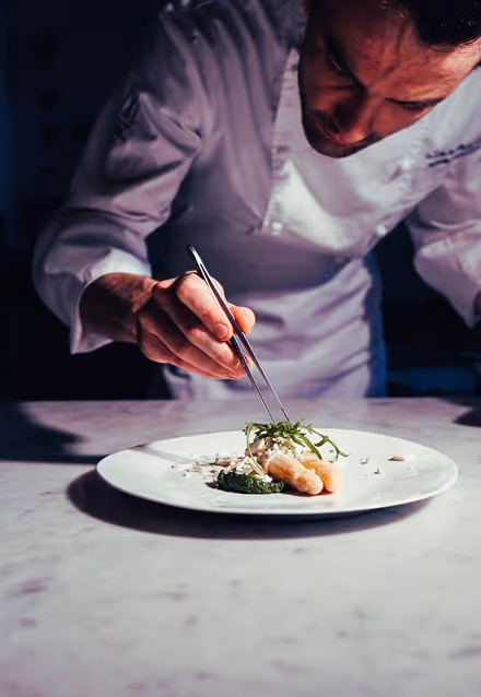 Chef in a white coat using tweezers to carefully garnish a plated dish on a white plate.