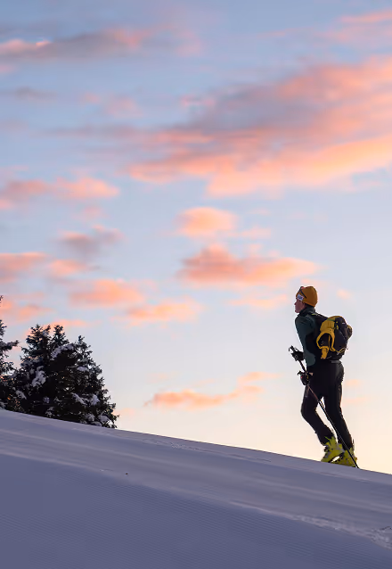 Person hiking uphill in snow with ski poles during a colorful sunset sky.