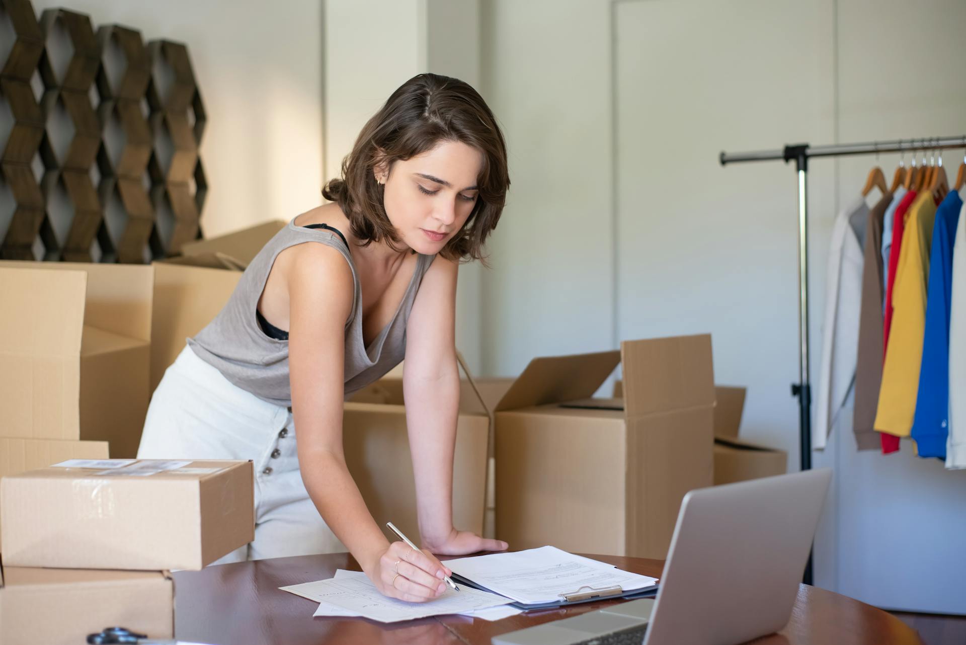 A young woman with dark hair, wearing a gray tank top and white skirt, is leaning over a wooden table to write on a document or checklist. She is surrounded by stacked cardboard boxes, suggesting she is preparing for a move or managing an e-commerce busine