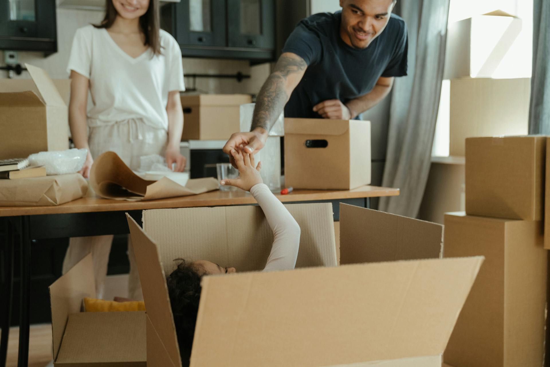 A lighthearted scene of a family during a move. A man is leaning over a large, open cardboard box and playfully holding the hand of a child lying inside it. A smiling woman is standing in the background next to a table covered with packing paper and other 