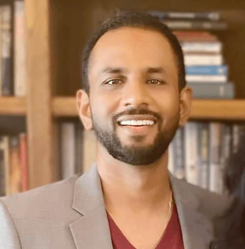 Smiling man with a short beard wearing a light gray blazer and maroon shirt standing in front of a bookshelf.