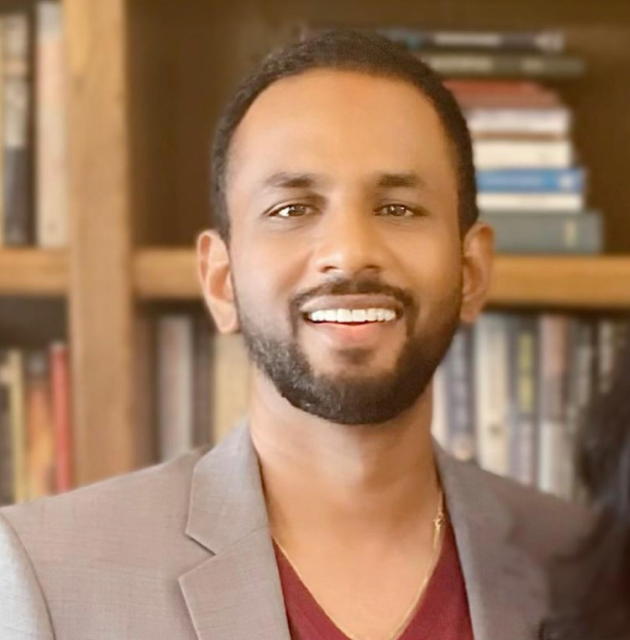 Smiling man with a short beard wearing a light gray blazer and maroon shirt standing in front of a bookshelf.