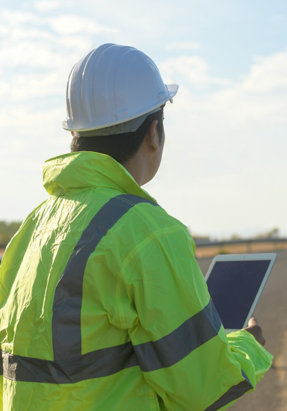 Construction worker in a white hard hat and neon yellow reflective jacket holding a tablet outdoors.