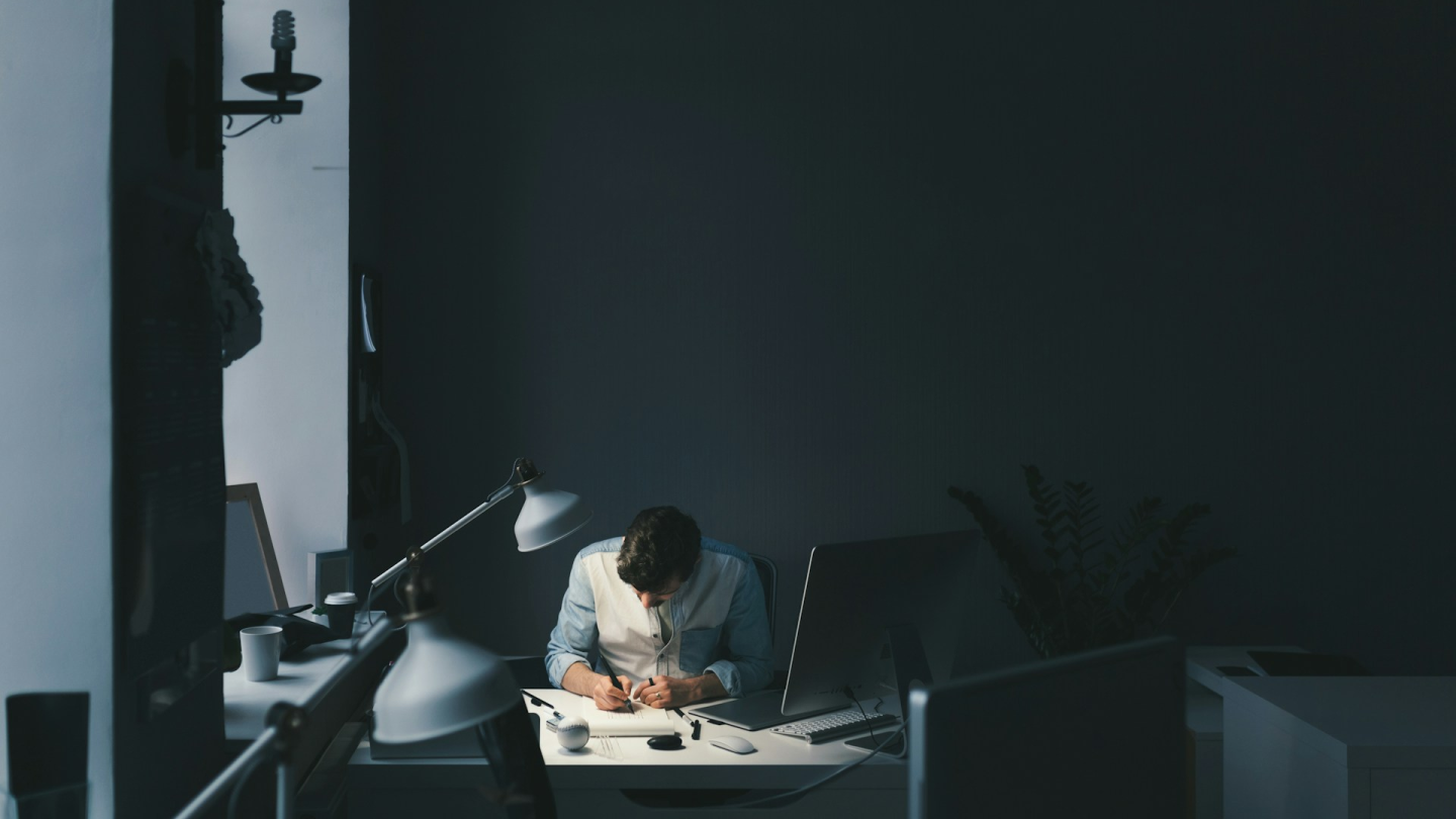 Man working late at a desk in a dimly lit room, writing on paper with a computer monitor and desk lamp nearby.