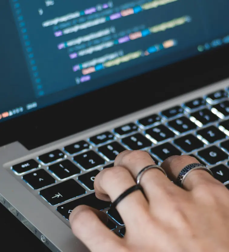 Hand typing on a backlit laptop keyboard with colorful lines of code on screen.