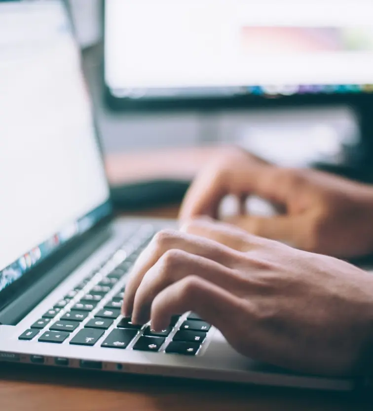 Close-up of hands typing on a laptop keyboard with a blurred monitor in the background.