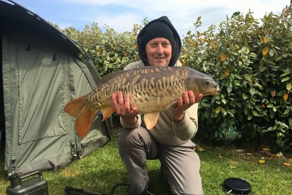 Man wearing a black beanie and hoodie holding a large fish while kneeling on grass near a tent and bushes.