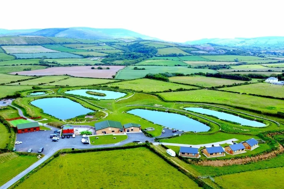Aerial view of a countryside fishing resort with multiple ponds, green fields, buildings, and parked cars surrounded by rolling hills.