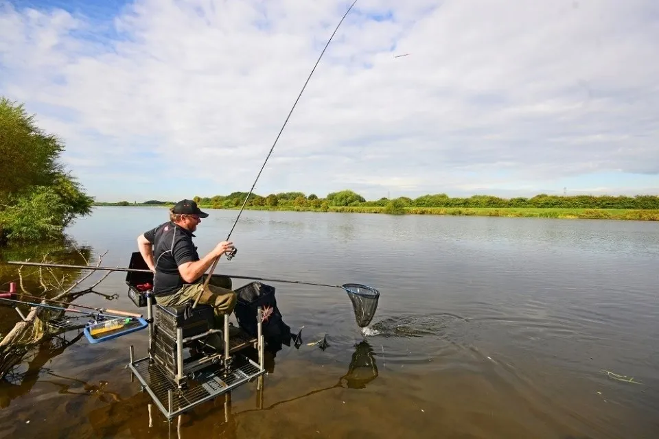 Man sitting on a fishing platform by a calm river, reeling in a fish with a net in the water.