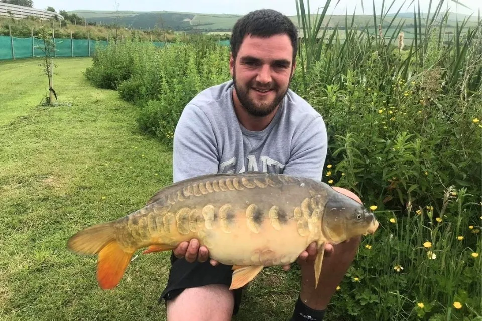 A man kneeling on grass holding a large mirror carp fish with greenery and hills in the background.