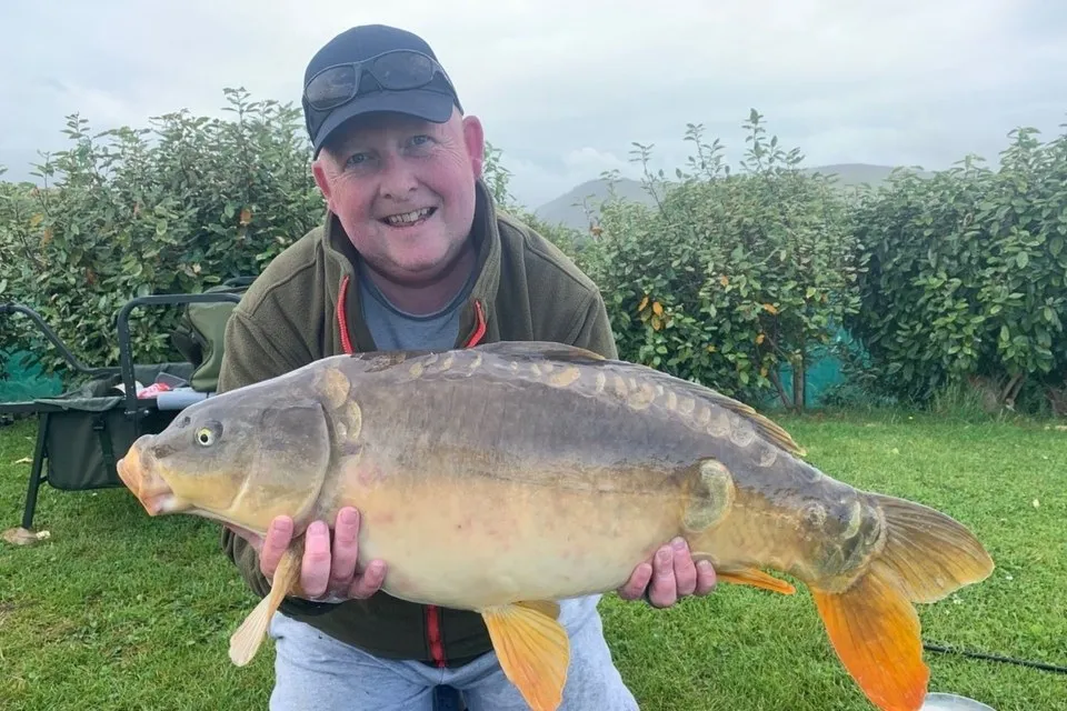 Man wearing a cap and jacket holding a large carp fish in front of green bushes and grass.