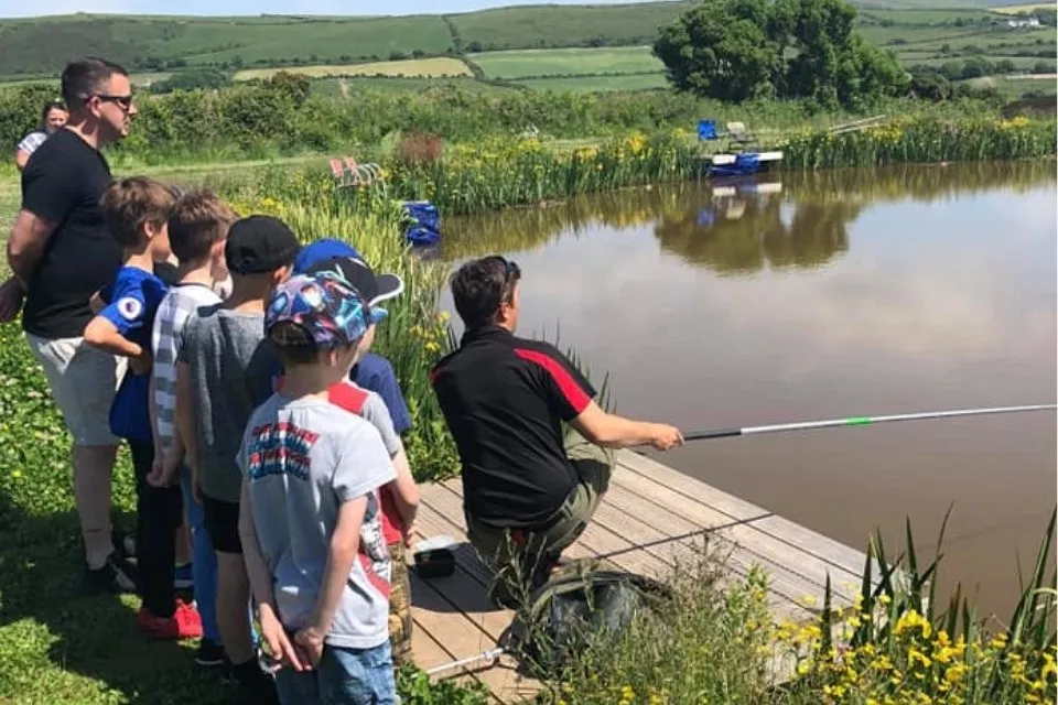 Man fishing on a wooden dock by a pond while a group of children and another adult watch.