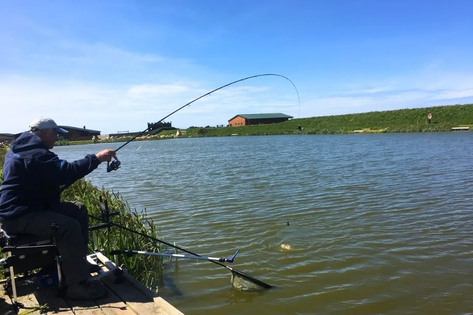 Man fishing by a lake, reeling in a fish near a wooden pier under a clear blue sky.