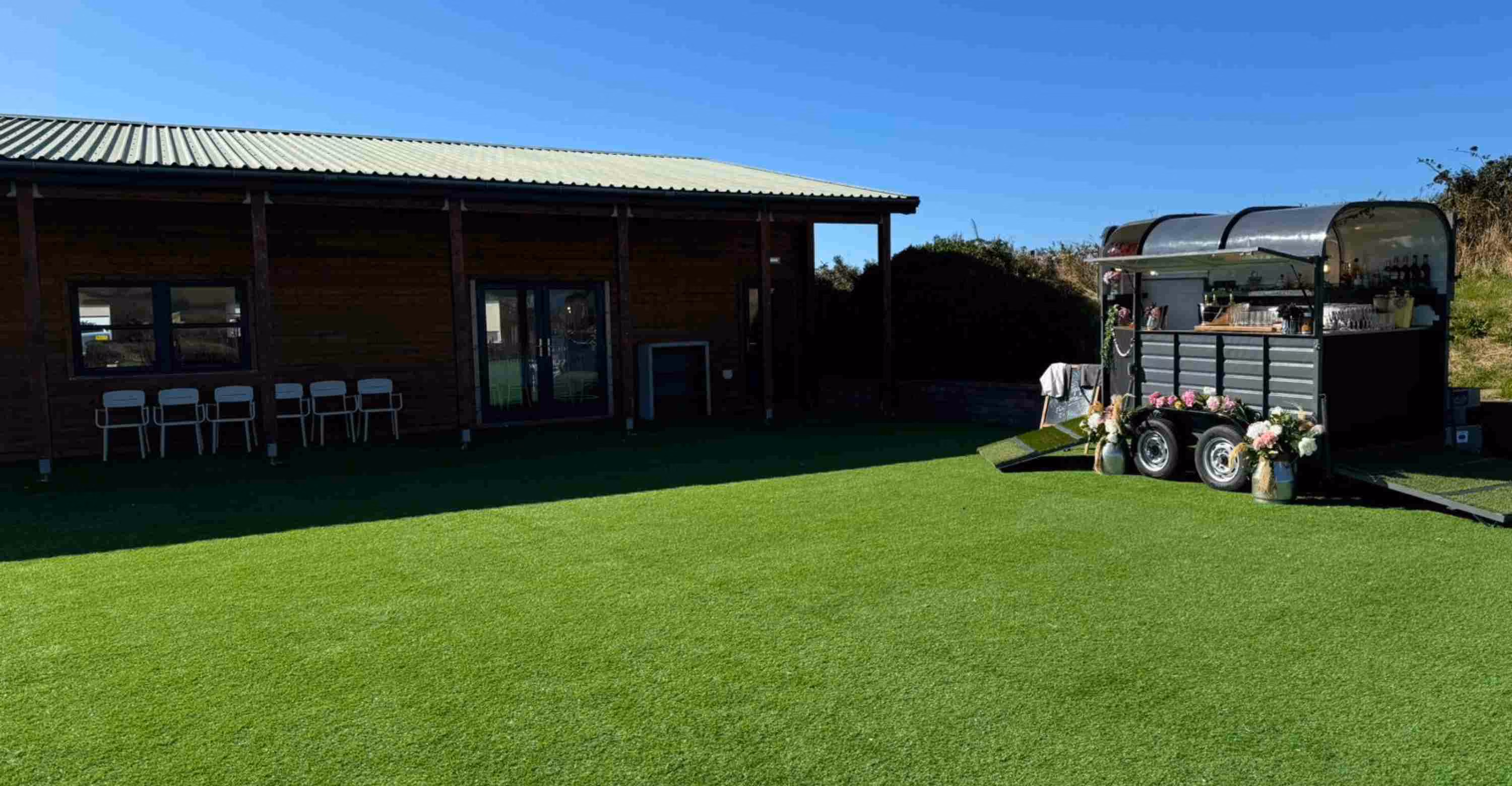 Outdoor event space with a green lawn, wooden building, and a decorated mobile bar trailer.