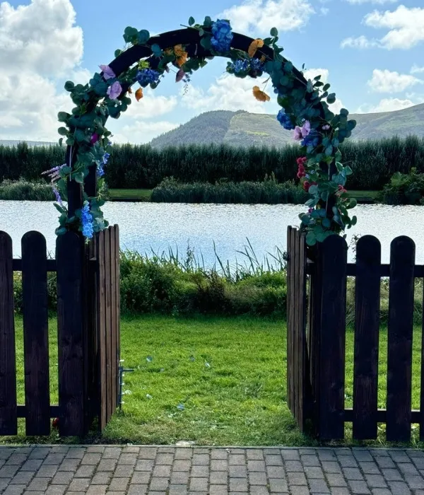 Wooden garden gate decorated with green leaves and blue and purple flowers, opening to a grassy area beside a lake with hills in the background.