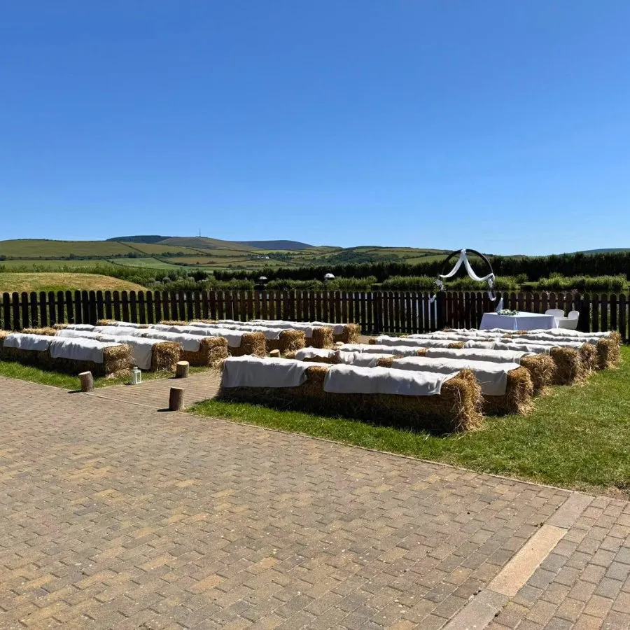 Outdoor wedding seating with hay bale benches covered in white cloths arranged on grass, with a wooden fence and scenic hills under a clear blue sky.