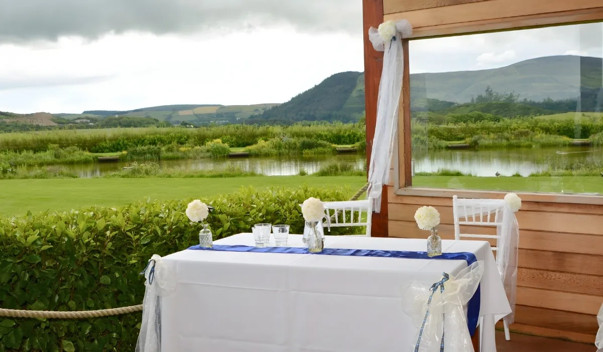 Wedding table with white cloth, blue runner, and white flower decorations near a wooden structure overlooking green fields and a pond under a cloudy sky.
