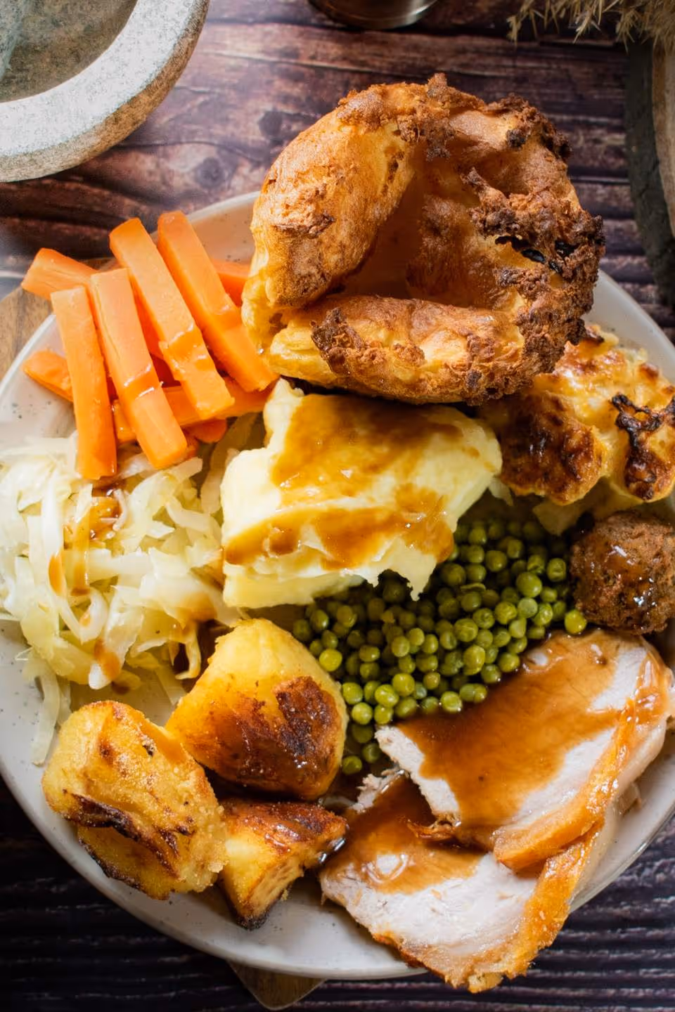 Plate with Sunday roast including roast potatoes, sliced meat with gravy, green peas, mashed potatoes with gravy, carrots, cabbage, Yorkshire pudding, and a meatball.