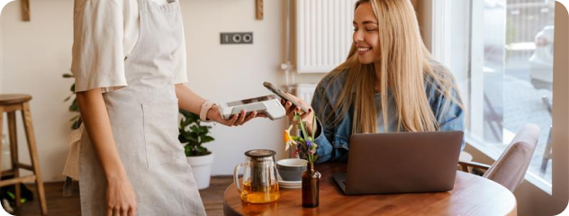Smiling woman with laptop paying via mobile phone to a cashier holding a card reader in a café.