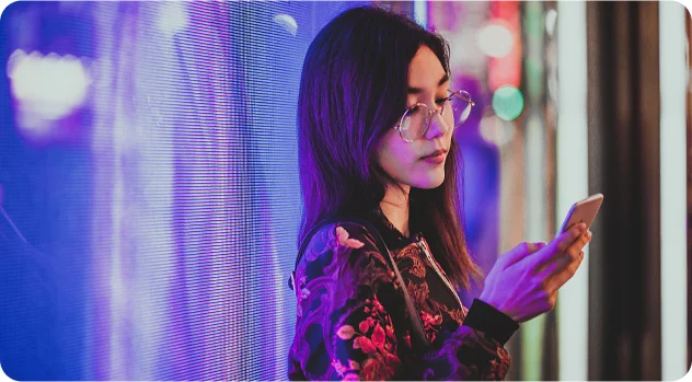 Young woman wearing glasses and a floral jacket looking at her smartphone with colorful blurry lights in the background.