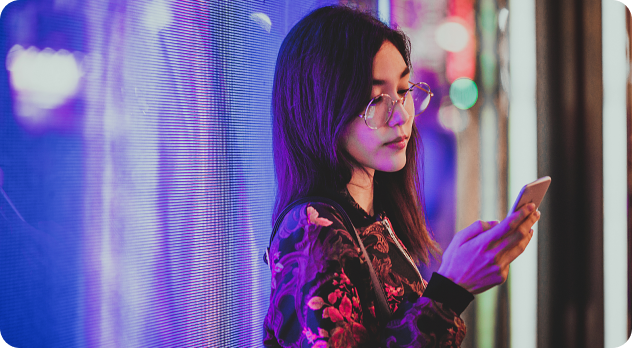 Young woman with glasses standing near a colorful LED-lit wall, looking at her smartphone.