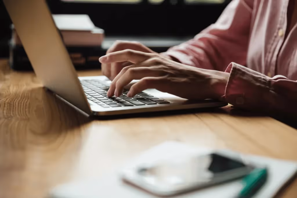 Close-up of someone typing on a laptop at a desk with a smartphone, books, and a pen.