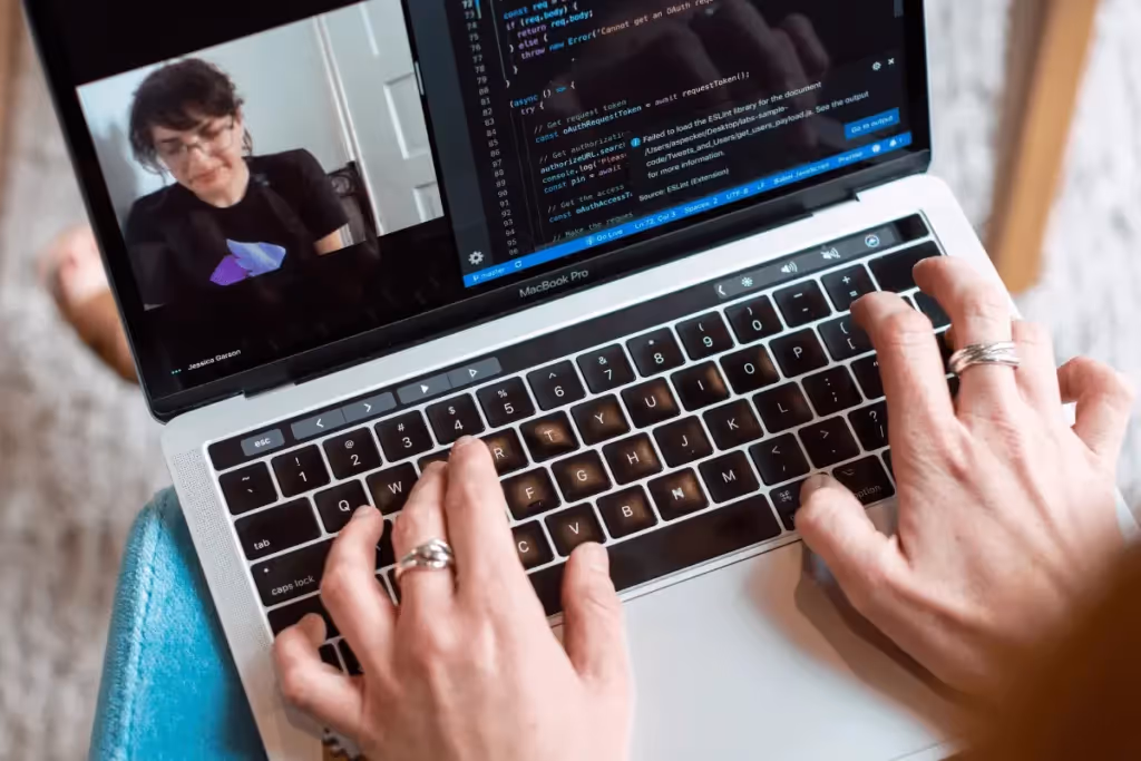 Hands typing on a laptop keyboard with a video call and code on the screen.