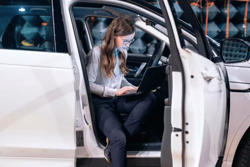 Woman sitting in a car, working on a laptop, performing diagnostics or programming.