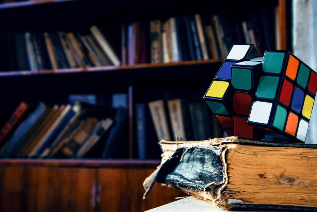 A colorful Rubik's Cube placed on an aged book with a library bookshelf in the background.