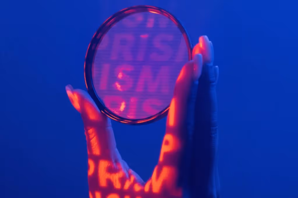 A close-up of a hand holding a circular lens, illuminated with red and blue neon lights, casting bold "PRISM" text projections.