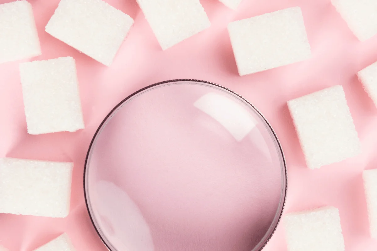 Magnifying glass surrounded by white sugar cubes on a soft pink background in a minimalist flat lay.