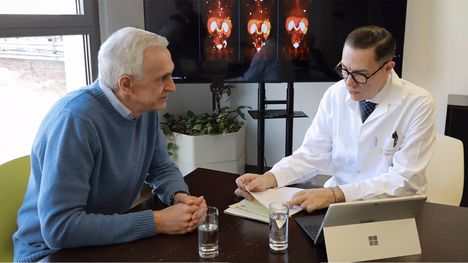 Dr. Marcus Hacker explains PSMA PET/CT imaging findings to an older patient with gray hair wearing a blue sweater, using medical images displayed on a monitor.