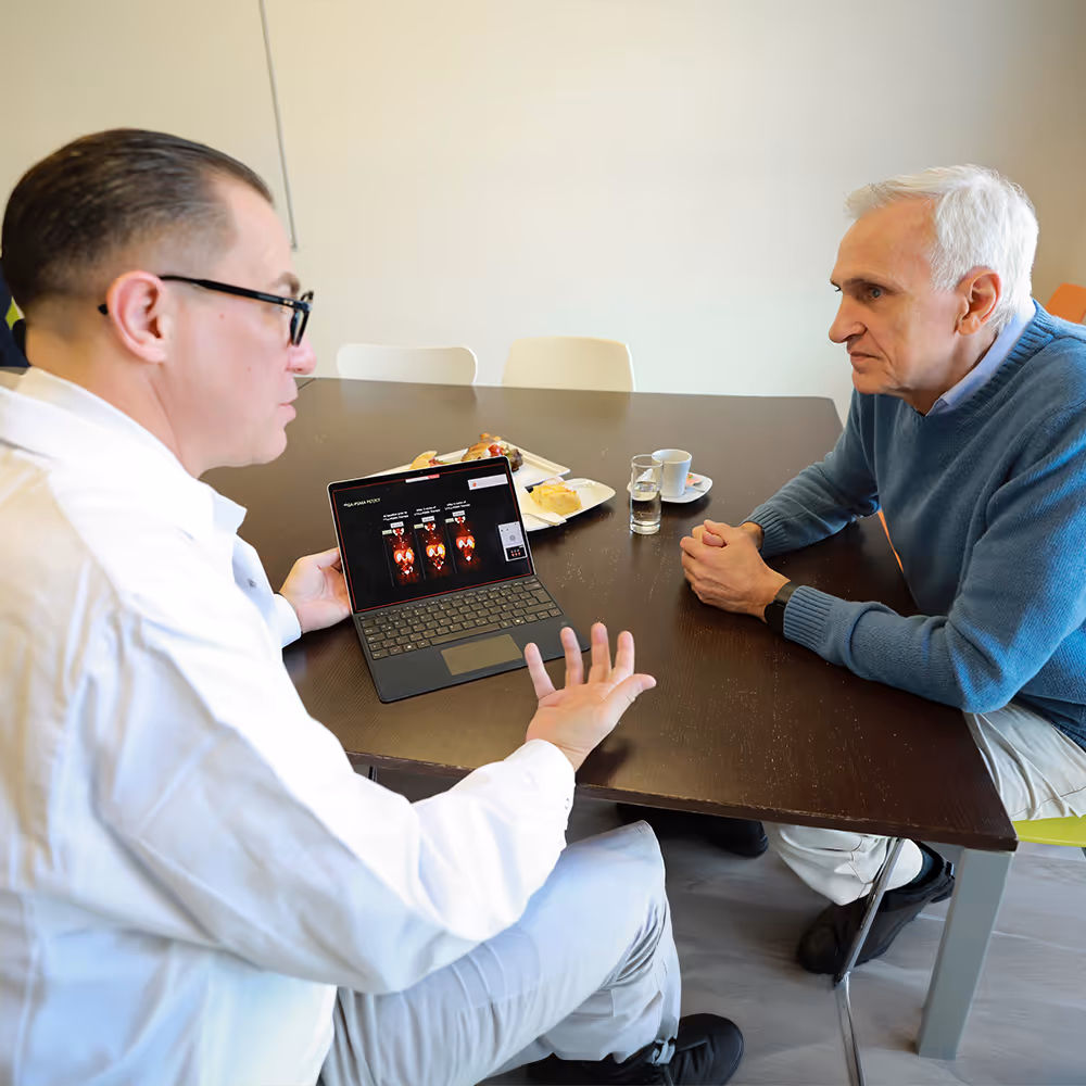 Dr. Marcus Hacker explains a PSMA PET/CT finding to an older patient wearing a blue sweater, using a laptop at a table.