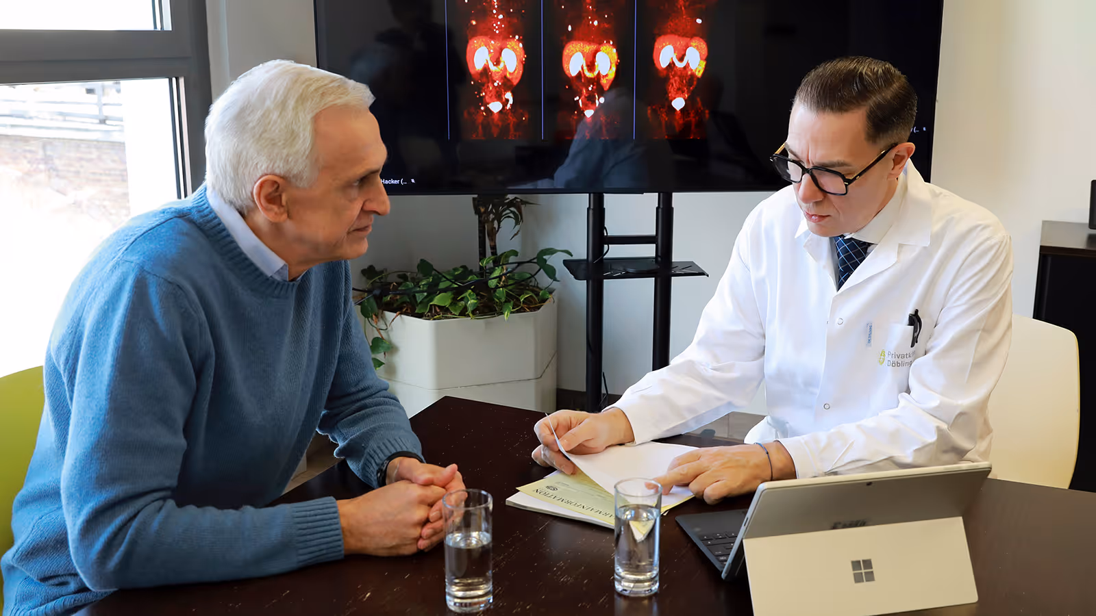 Dr. Marcus Hacker erklärt einem älteren Patienten mit grauem Haar und blauem Pullover medizinische Bilder eines PSMA-PET/CT-Befundes auf einem Monitor.