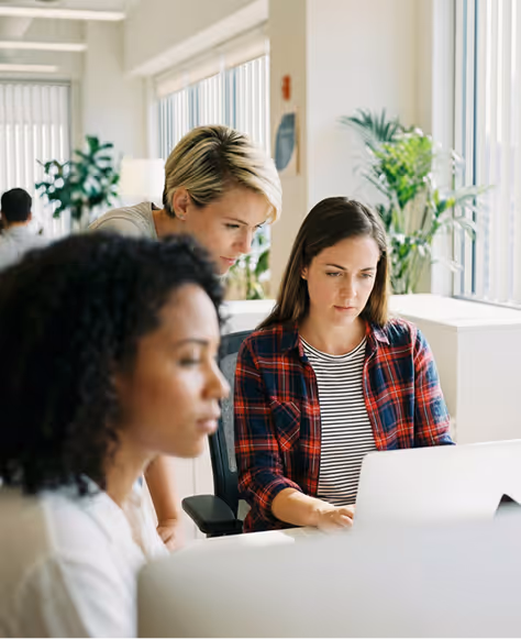 Three women working together in a bright office with one woman looking at a laptop screen while another leans over her shoulder.