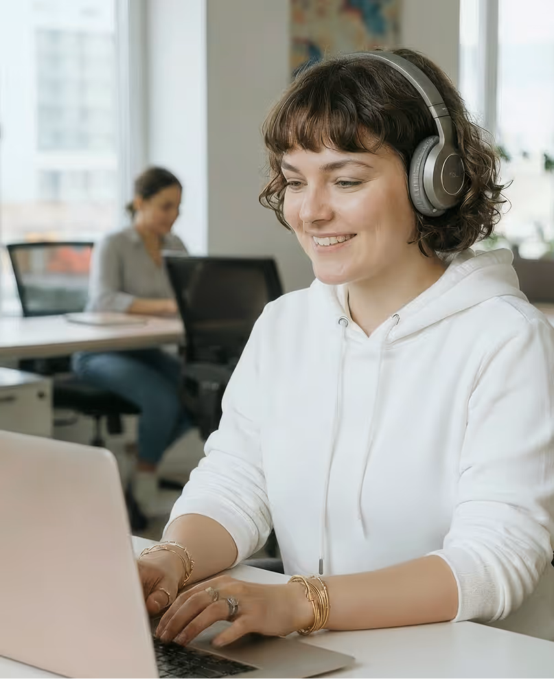 Smiling woman wearing headphones and a white hoodie typing on a laptop in an office with another person working in the background.