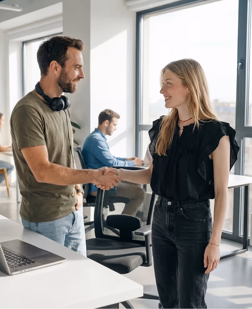 Man and woman smiling and shaking hands in a bright modern office with coworkers in the background.