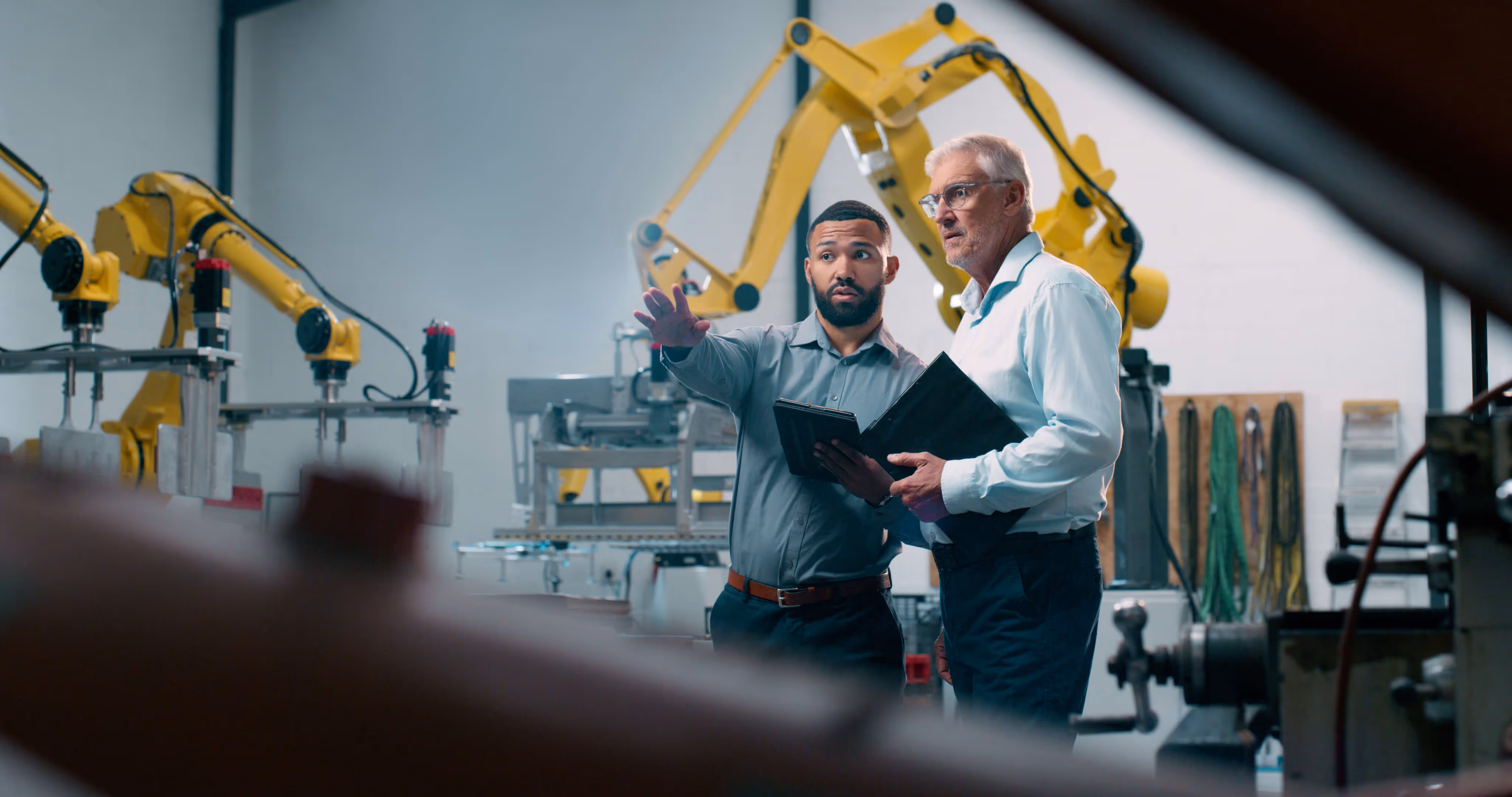 Two men discussing and pointing in a factory with yellow robotic arms in the background.