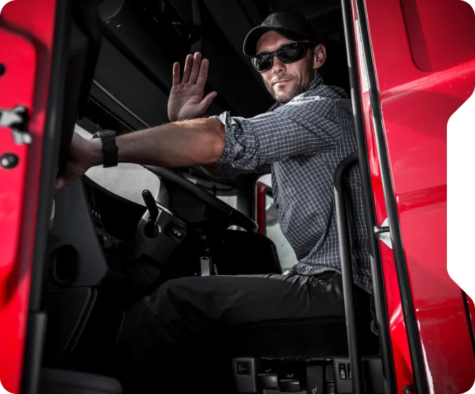 Man wearing sunglasses and a cap sitting in the driver's seat of a red truck, waving with his right hand.