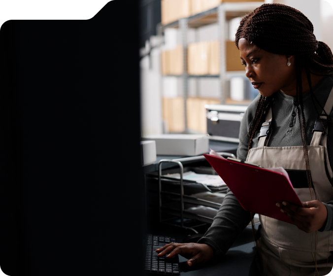 Woman in overalls holding a red clipboard typing on a keyboard in a storage room with shelves filled with boxes.
